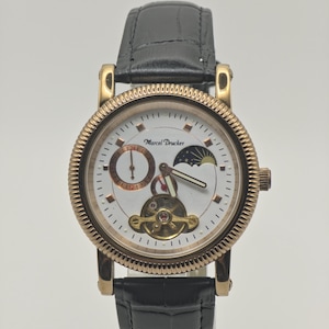 May include: A Marcel Drucker wristwatch with a gold-tone fluted bezel and a white dial. The watch features a black leather strap with a crocodile texture, a moon phase indicator, a 24-hour sub-dial, and an open heart balance wheel movement.