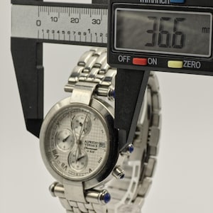May include: A silver Alfredo Versace chronograph watch with a stainless steel band. The watch face has three sub-dials and Roman numeral hour markers. The watch measures 36.6 mm in diameter.
