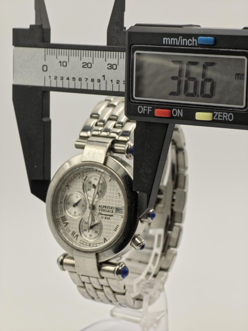 May include: A silver Alfredo Versace chronograph watch with a stainless steel band. The watch face has three sub-dials and Roman numeral hour markers. The watch measures 36.6 mm in diameter.