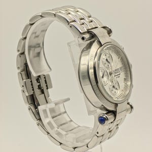 May include: A silver-toned wristwatch with a textured white face and multiple sub-dials. The watch features a stainless steel band and a blue gem accent. The watch is displayed on a clear stand.