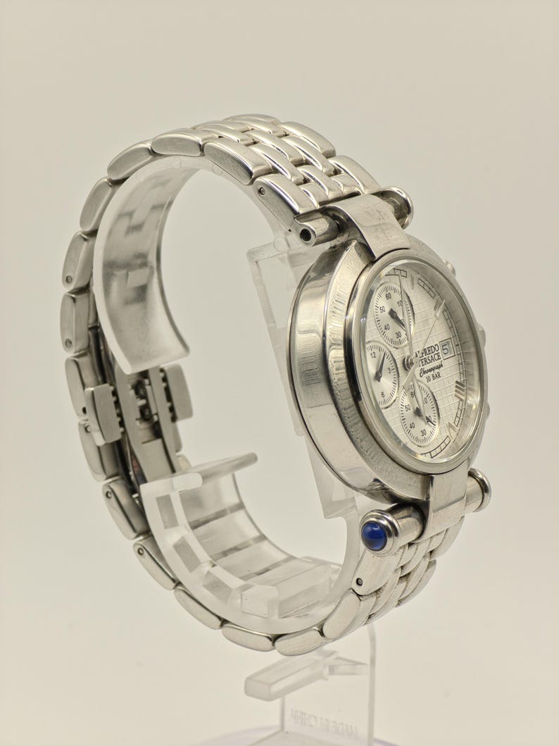 May include: A silver-toned wristwatch with a textured white face and multiple sub-dials. The watch features a stainless steel band and a blue gem accent. The watch is displayed on a clear stand.