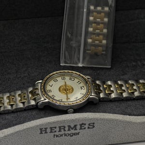 May include: A silver and gold-toned wristwatch with a white face and gold accents. The watch has a silver band with gold-toned links. The watch is displayed on a gray surface with the brand name "HERMES" and the word "horloger" printed on it.