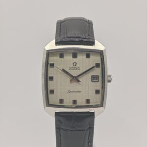 Puede incluir: Un reloj de pulsera automático Omega Seamaster vintage con esfera plateada y ventana de fecha a las 3. El reloj tiene una caja rectangular de acero inoxidable y una correa de cuero negro. La esfera presenta marcadores de hora cuadrados negros.