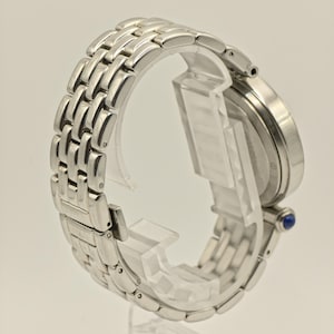 May include: A silver wristwatch with a linked band is displayed on a clear stand. The watch face is round, and a small blue gem is visible on the side. The band is made of multiple rectangular links.