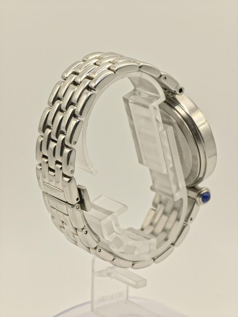 May include: A silver wristwatch with a linked band is displayed on a clear stand. The watch face is round, and a small blue gem is visible on the side. The band is made of multiple rectangular links.