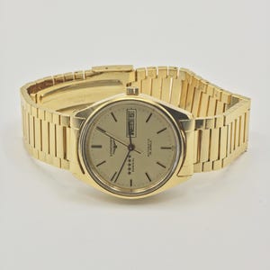 May include: A gold-toned Longines Admiral automatic wristwatch. The watch has a gold dial with black hour markers and hands, a day-date display, and a gold-toned bracelet. The watch is displayed on a neutral background.