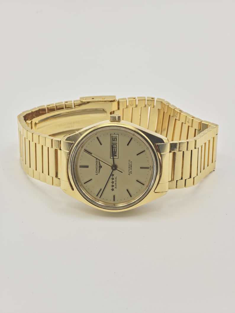 May include: A gold-toned Longines Admiral automatic wristwatch. The watch has a gold dial with black hour markers and hands, a day-date display, and a gold-toned bracelet. The watch is displayed on a neutral background.