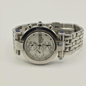 May include: A silver-toned wristwatch with a round face and a linked band. The watch face has multiple sub-dials and Roman numeral hour markers. The watch has blue accents and the brand name "ALFREDO VERSACE" is visible.
