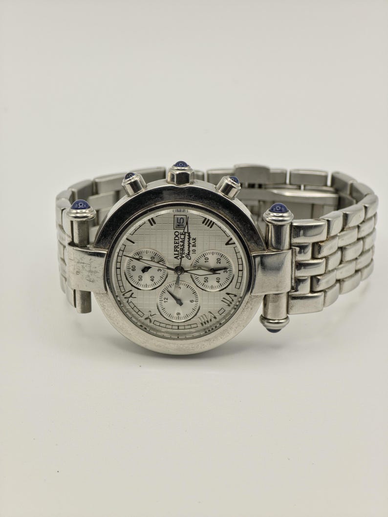 May include: A silver-toned wristwatch with a round face and a linked band. The watch face has multiple sub-dials and Roman numeral hour markers. The watch has blue accents and the brand name "ALFREDO VERSACE" is visible.