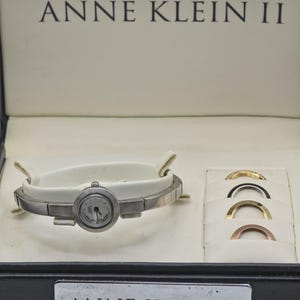 May include: An Anne Klein II watch set in a presentation box. The silver-tone watch features a round face and a white band. Four interchangeable metal bezels in gold, silver, rose gold, and black are included in the box, which displays the brand name.