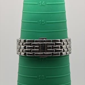 May include: A silver-toned metal watch band with a rectangular link design. The band is clasped and resting on a green ring sizer. The sizer has measurements from 14 to 19.