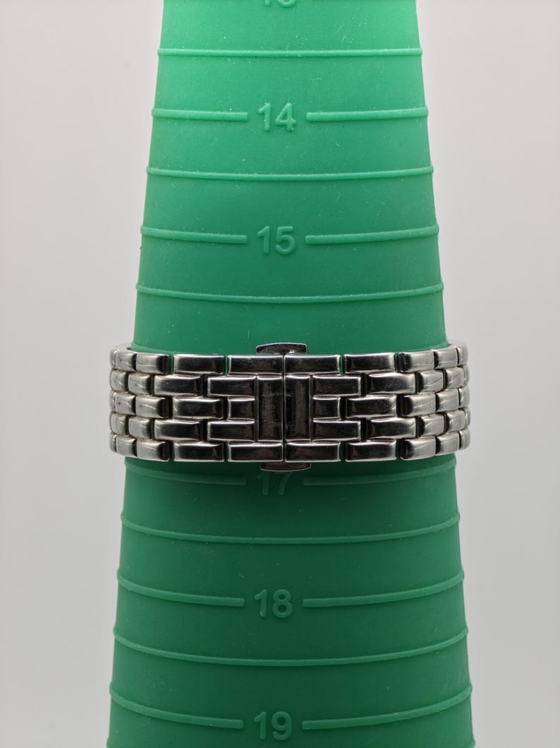 May include: A silver-toned metal watch band with a rectangular link design. The band is clasped and resting on a green ring sizer. The sizer has measurements from 14 to 19.