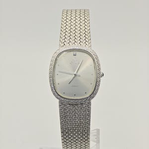 Puede incluir: Un reloj de cuarzo Cyma plateado con esfera ovalada, fondo plateado texturizado y un diamante único en la posición de las doce. El reloj cuenta con una correa de malla metálica tejida y un borde texturizado a juego alrededor de la caja del reloj.