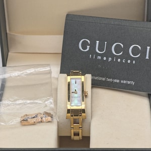 Puede incluir: Un reloj Gucci Timepieces dorado con esfera rectangular y correa a juego, presentado en su caja original. La esfera del reloj tiene un aspecto nacarado. La caja incluye una tarjeta de garantía y el logotipo de Gucci.