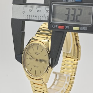May include: A gold Longines Admiral automatic wristwatch. The watch face shows the day and date, with the text "WED 5". The watch is measured with a digital caliper, showing a width of 33.2 mm.