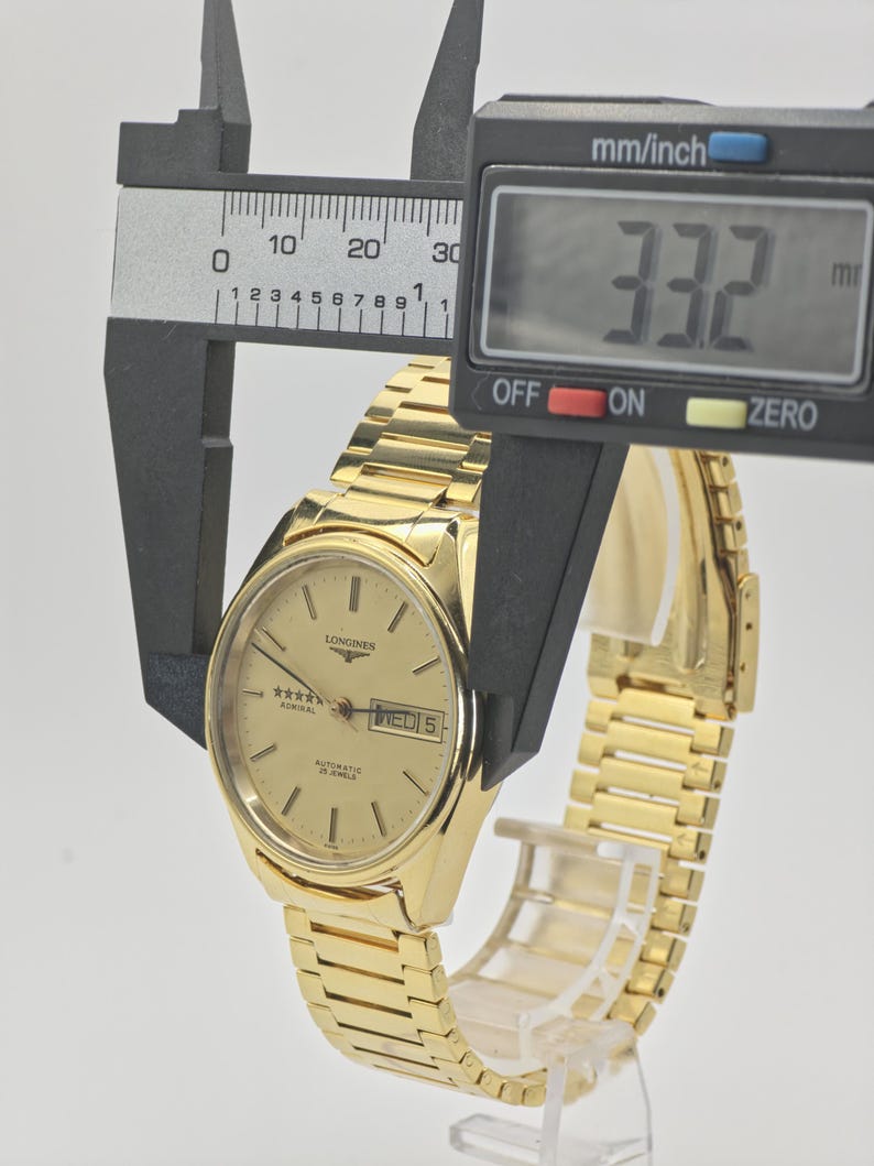 May include: A gold Longines Admiral automatic wristwatch. The watch face shows the day and date, with the text "WED 5". The watch is measured with a digital caliper, showing a width of 33.2 mm.