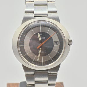 Könnte beinhalten: Eine silberfarbene Omega Genève Dynamic Automatik-Armbanduhr. Die Uhr hat ein dunkelgraues Zifferblatt mit orangefarbenen Zeigern und Datumsfenster. Das Armband ist aus Edelstahl.