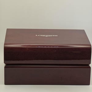 May include: A rectangular, glossy, dark burgundy watch box. The top of the box has the word "LONGINES" in silver lettering. The box is made of wood and has a two-piece design with a hinged lid.