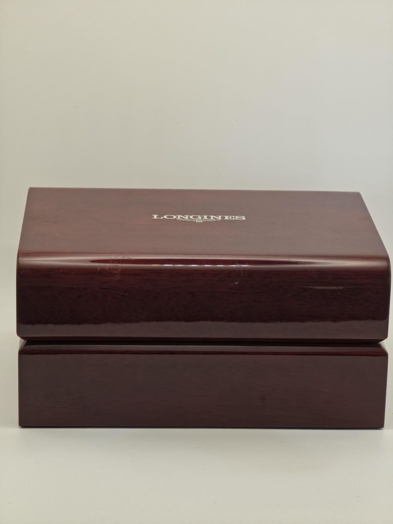 May include: A rectangular, glossy, dark burgundy watch box. The top of the box has the word "LONGINES" in silver lettering. The box is made of wood and has a two-piece design with a hinged lid.