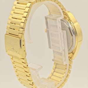 May include: A gold wristwatch with a rectangular face and a matching gold link strap. The watch is displayed on a clear plastic stand, highlighting the detailed design of the strap and the watch's overall appearance. The strap is made of small rectangular links.