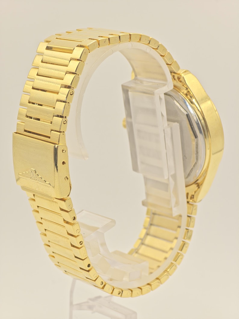 May include: A gold wristwatch with a rectangular face and a matching gold link strap. The watch is displayed on a clear plastic stand, highlighting the detailed design of the strap and the watch's overall appearance. The strap is made of small rectangular links.