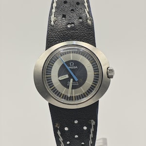 May include: A vintage Omega Dynamic wristwatch with a black leather strap. The watch face is silver and black with the Omega logo and the words "Dynamic" and "Geneve". The strap has white stitching and perforations.