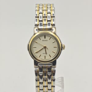 Puede incluir: Un reloj de pulsera Seiko Lucent con esfera redonda de color crema, marcadores de hora y manecillas en tono dorado. El reloj cuenta con una correa de eslabones de acero inoxidable bicolor con segmentos plateados y dorados, y un bisel dorado.