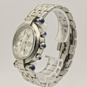 May include: A silver-toned Versace chronograph watch with a textured face and multiple sub-dials. The watch features a stainless steel band and is accented with blue gemstones. The watch is displayed on a clear stand.
