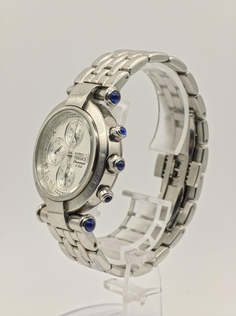 May include: A silver-toned Versace chronograph watch with a textured face and multiple sub-dials. The watch features a stainless steel band and is accented with blue gemstones. The watch is displayed on a clear stand.