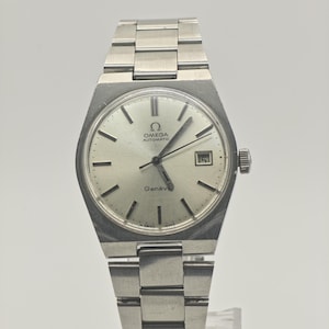 May include: Silver Omega Genève automatic wristwatch with a silver dial and a date window at the 3 o'clock position. The watch has a stainless steel bracelet and a round face. The watch is displayed on a clear stand.
