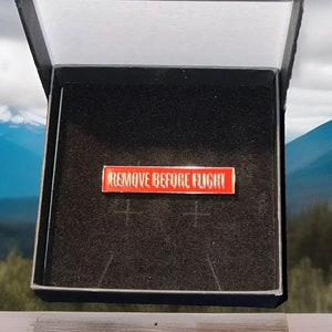 以下が含まれることがあります： 白い文字で「REMOVE BEFORE FLIGHT」と書かれた赤い長方形のピン。ピンは、白い内側の黒いベルベットの箱に入っています。