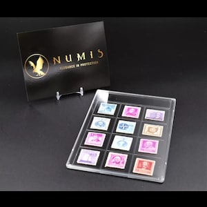 May include: A clear plastic display case holding twelve colorful stamps. A black card with gold text and a logo that reads "NUMIS Elegance in Protection" is propped up behind the case. The stamps are in various colors and feature different designs.
