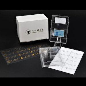 May include: A collection of numismatic supplies, including a white box with the "NUMIS" logo, a graded stamp in a clear holder, and various packaging materials. The stamp is light blue with a plane.