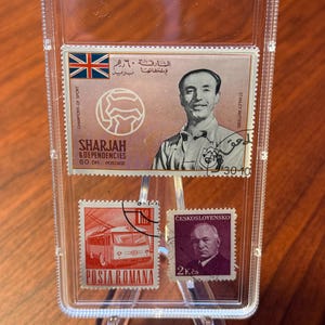 May include: A clear plastic case holding four vintage postage stamps. The stamps feature images of a soccer player, a trolley, and portraits. The top stamp has the Union Jack and Arabic text. The stamps are displayed on a wooden surface.