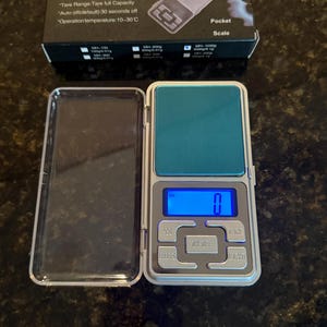 Puede incluir: Una báscula de bolsillo plateada con una pantalla azul que muestra "0". La báscula tiene botones etiquetados como "PCS", "UNITS", "TARE" y "LIGHT". La caja dice "Pocket Scale" y enumera las características estándar.