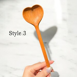 May include: A wooden spoon shaped like a heart. The spoon is brown and has a smooth finish. The spoon is held in a hand.