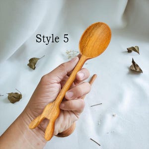 May include: A wooden spoon with a unique bunny-shaped handle. The spoon is made of natural wood and has a smooth, polished finish. The spoon is held in a hand, and the background is a white surface.