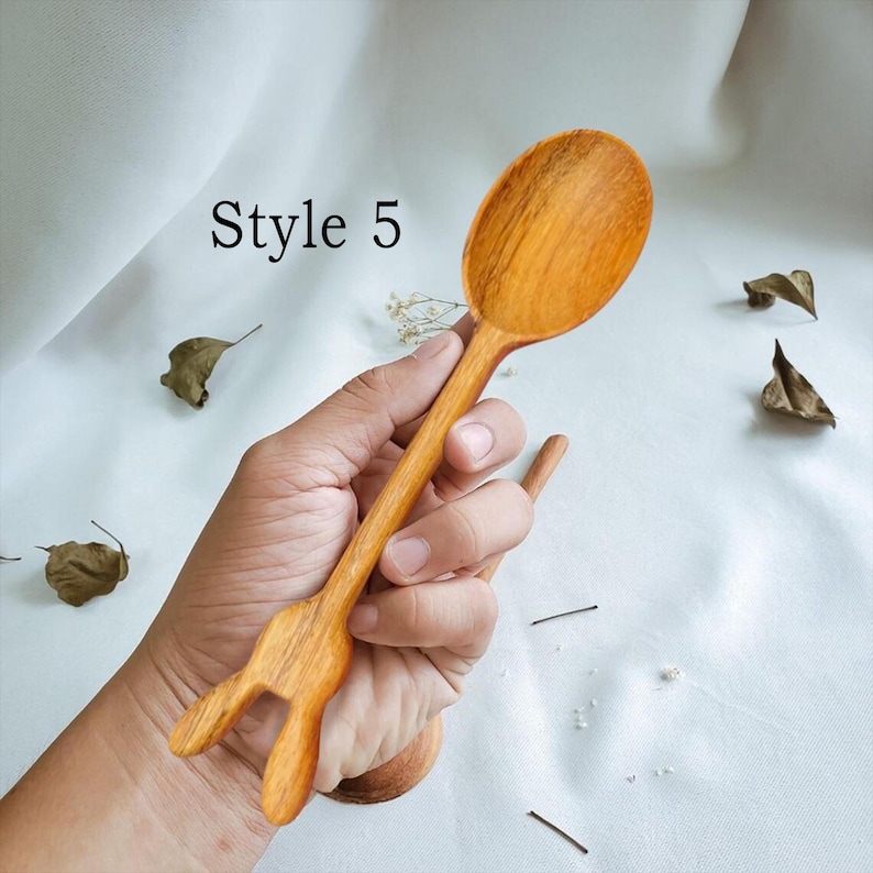 May include: A wooden spoon with a unique bunny-shaped handle. The spoon is made of natural wood and has a smooth, polished finish. The spoon is held in a hand, and the background is a white surface.
