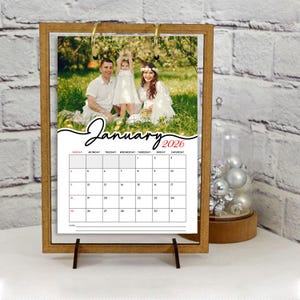 May include: A wooden framed calendar for January 2026, featuring a family photo. The calendar has a simple grid layout with the days of the week and dates. The frame is brown and the calendar is propped up on a stand.
