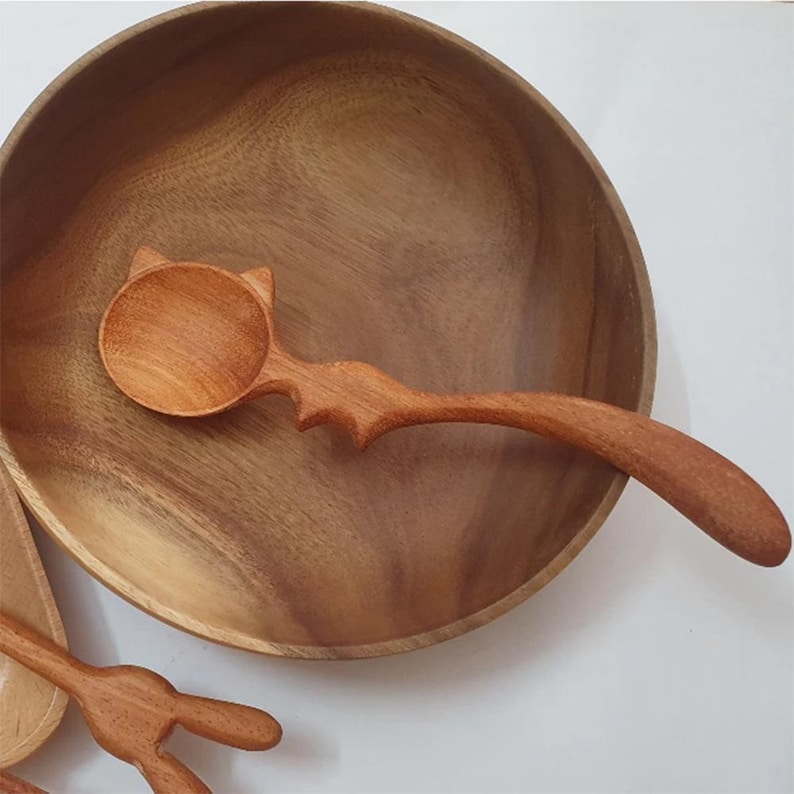May include: A wooden spoon with a cat-shaped handle, resting inside a wooden bowl. The spoon is made of natural wood and has a smooth, rounded surface.