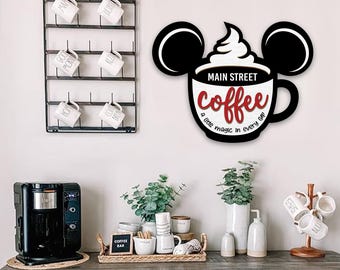 May include: A wooden coffee cup-shaped sign with black mouse ears, the words "Main Street Coffee" and "a little magic in every cup" in red and white. The sign is mounted on a white wall above a coffee bar.