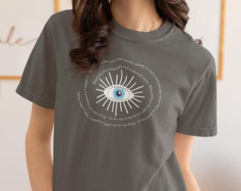 Puede incluir: Camiseta gris oscuro con un diseño de ojo blanco y azul y texto. El ojo tiene un iris azul y está rodeado de líneas radiantes. El texto añade un elemento espiritual o místico al diseño, ideal para un look casual.