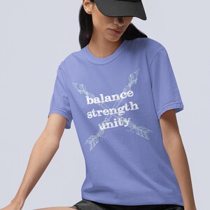 Può includere: T-shirt lilla chiaro con le parole bianche "balance strength unity". Due frecce si incrociano dietro il testo. La maglietta ha girocollo e maniche corte.