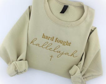 May include: A beige sweatshirt with the text "hard fought hallelujah" embroidered in gold with a small gold cross below the text.