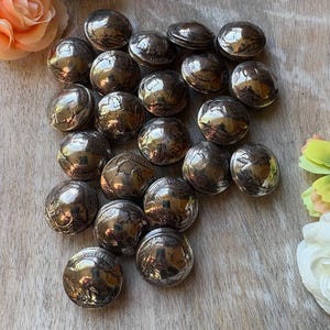 May include: A collection of silver-colored, coin-shaped objects, possibly buttons or decorative elements. The objects are arranged on a light-colored, textured surface, with floral accents in the corners. The coins have detailed designs and reflect light.