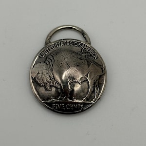 Puede incluir: Colgante plateado con un diseño de búfalo, con las inscripciones "UNITED STATES OF AMERICA" y "FIVE CENTS". El colgante tiene un lazo en la parte superior para sujetarlo a una cadena o cordón.
