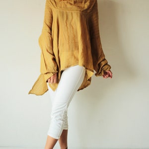 May include: A mustard yellow, loose-fitting, long-sleeved top with a draped neckline and a high-low hemline. The top is made of a lightweight fabric and has a relaxed fit. The model is wearing white cropped pants and black heeled sandals.