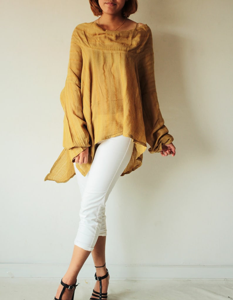 May include: A mustard yellow, loose-fitting, long-sleeved top with a draped neckline and a high-low hemline. The top is made of a lightweight fabric and has a relaxed fit. The model is wearing white cropped pants and black heeled sandals.