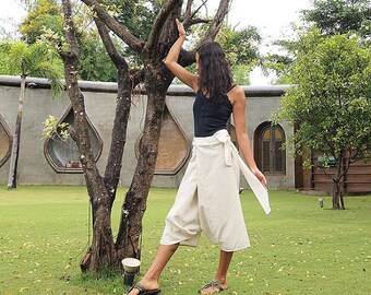 May include: A woman wearing a black tank top and cream-colored wrap pants. The pants are tied at the waist with a long sash. She is standing near a tree in a grassy area, wearing sandals.