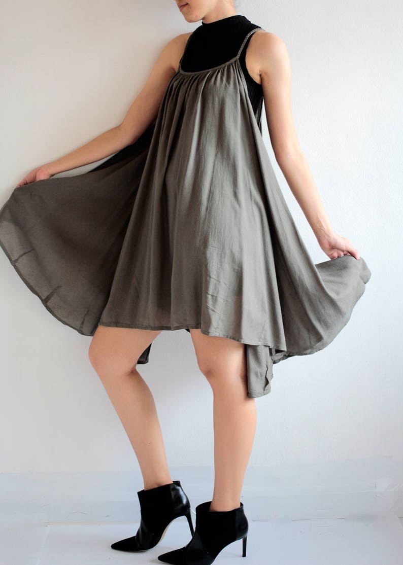 May include: A grey sleeveless dress with a loose, flowing fit. The dress has a high neckline and a slightly asymmetrical hemline. The model is wearing black ankle boots.